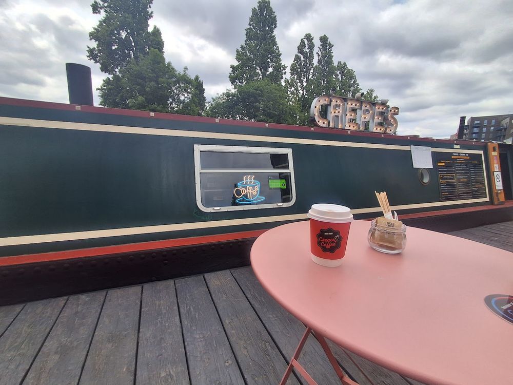 The green narrowboat with its serving hatch and a pink table on the deck, moored on the Grand Union canal